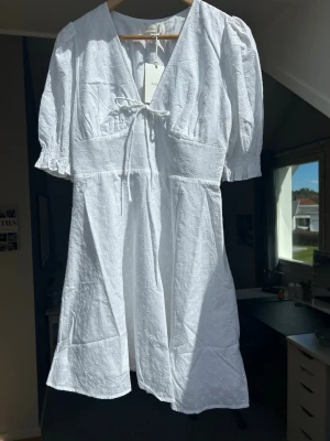 Vit kort klänning med puffärm - Söt vit kort klänning med broderade detaljer, puffärmar och v-ringning. Klänningen har knyt framtill och resår i midjan bak för en bekväm passform. Helt nyköpt för 899 kr, men säljer pga att jag redan har en klänning till skolavslutningen. 