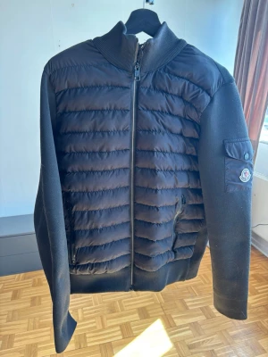 Svart pufferjacka från Moncler - Snygg svart pufferjacka från Moncler med quiltad front, ribbade muddar och ärmar i mjukt material. Jackan har dragkedja framtill, fickor med dragkedja och en ikonisk Moncler-logga på ärmen. Perfekt för kalla vinterdagar och riktigt stilren look.