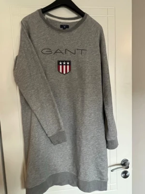 Grå sweatshirtklänning från GANT - Grå sweatshirtklänning från GANT med broderad logga och sköld på bröstet. Klänningen har rund halsringning, långa ärmar och är gjord i mjukt sweatshirtmaterial för en chill och sportig vibe.