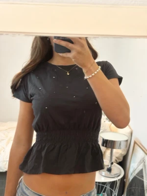 Svart topp med pärlor och resårmidja - Snygg svart topp med korta ärmar och rund hals. Toppen har små pärlor framtill och en smockad resår i midjan som ger en fin form. Tillverkad i mjuk bomull och passar perfekt till jeans eller kjol.
