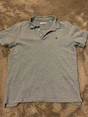 Ljusblå pikétröja från Jack & Jones - Snygg ljusblå pikétröja från Jack & Jones med klassisk krage och knappar framtill. Tröjan har en liten broderad logga på bröstet och är tillverkad i mjuk bomull. Perfekt för en avslappnad och stilren look. (Liten i storleken)