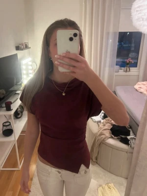 Vinröd topp med asymmetrisk nederkant - Snygg vinröd topp med korta ärmar och unik asymmetrisk nederkant. Toppen har en figurnära passform och är tillverkad i ett mjukt, stretchigt material som sitter bekvämt. Perfekt att styla med jeans eller kjol för en trendig look.