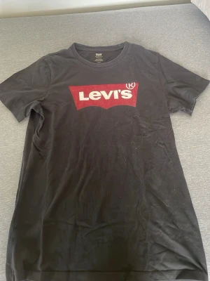 Svart Levi's t-shirt med logga - Cool svart t-shirt från Levi's med klassisk röd och vit logga på bröstet. T-shirten har rund hals och korta ärmar, perfekt för en avslappnad och stilren look. Materialet är mjuk bomull som känns skönt mot huden.