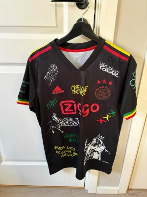 Ajax x Bob Marley limited edition tröja - Säljer en svart Ajax x Bob Marley limited edition matchtröja från Adidas. Tröjan har färgglada detaljer i rött, gult och grönt, tryck med Bob Marley, citat och Ajax-logga. Materialet är lätt och andas, perfekt för fotboll eller streetwear.