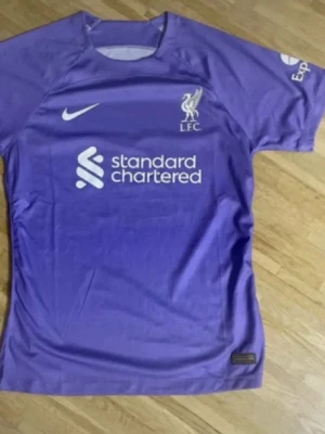 Lila Liverpool Nike matchtröja - Snygg lila matchtröja från Liverpool FC med Nike-logga och Standard Chartered sponsortryck på bröstet. Tröjan har korta ärmar och klubbmärke på vänster sida. Perfekt för dig som älskar fotboll och vill visa ditt stöd för LFC.