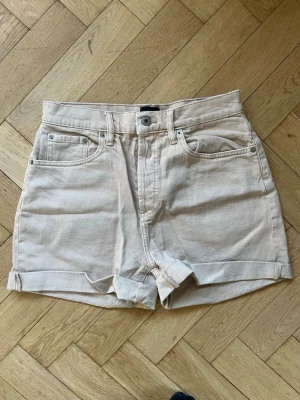 Beige högmidjade jeansshorts - Säljer ett par beige jeansshorts från Lager 157 med hög midja och klassisk femficksdesign. Shortsen har uppvikta benslut och är tillverkade i bomull, vilket gör dem både bekväma och snygga. I modellen Diba.