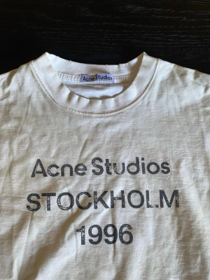 Vit Acne Studios t-shirt med tryck - Vit t-shirt från Acne Studios med svart texttryck 'STOCKHOLM 1996' på framsidan. Klassisk rund hals och korta ärmar, perfekt för dig som gillar minimalistisk och stilren design. Materialet är mjuk bomull som känns skönt mot huden.
