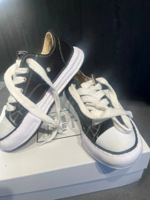 Maison miharas - Snygga svarta sneakers med vita tjocka skosnören och vit sula Maison miharas Klassisk låg modell med kontrastsömmar och rund tå. Ovandelen är i tyg och sulan i gummi. Perfekta för en avslappnad streetstil.