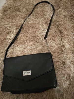 Svart axelväska från Guess - Snygg svart axelväska från Guess med justerbar axelrem och silverfärgad logga framtill. Väskan har lock med magnetknäppning och diskret mönster med GG-loggor på framsidan. Tillverkad i konstläder och har en stilren, rektangulär form.