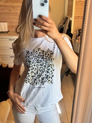 Vit t-shirt med leopardmönster - Säljer en vit t-shirt med stort leopardmönster framtill och textdetalj längst ner. T-shirten har korta ärmar och en avslappnad passform, perfekt för en cool och trendig look. Passar dig som gillar djurmönster och vill sticka ut lite extra.