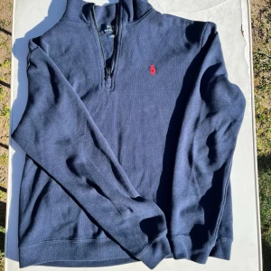 Marinblå half zip tröja från Polo Ralph Lauren - Klassisk marinblå tröja från Polo Ralph Lauren med half zip och röd broderad logga på bröstet. Tröjan har lång ärm och ribbade muddar vid ärmslut och nederkant. Perfekt för en stilren och avslappnad look.