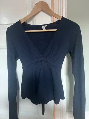 Mörkblå omlottopp med knyt - Mörkblå omlottopp med v-ringning och långa ärmar. Toppen har ett knytband i midjan som ger en snygg siluett och är tillverkad i ett ribbat material. Perfekt för dig som gillar stilrena och trendiga plagg.