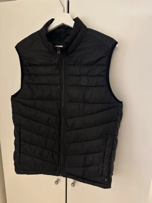 Jack & Jones Väst - Snygg svart väst från Jack & Jones. Västen har ett quiltat mönster och två sidofickor. Perfekt att slänga över en hoodie eller tröja för extra värme. Enkel och stilren design som passar till det mesta. Den har små hål nertill (se bild), samt ett pyttelitet hål på vänster sida men inget alls man tänker på.