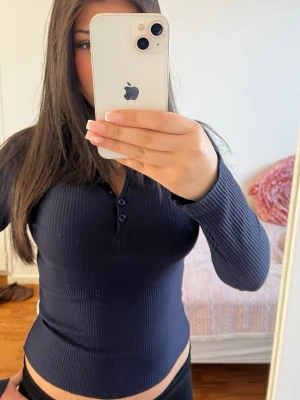 Mörkblå ribbad långärmad topp - Säljer en mörkblå ribbad långärmad topp med v-ringning och knappar framtill. Toppen har en slim passform och är tillverkad i ett stretchigt material som sitter snyggt på kroppen. Perfekt att styla med jeans eller kjol.