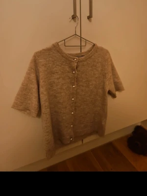 Beige stickad kofta med pärlknappar - Mysig beige stickad kofta med korta ärmar och pärlknappar framtill. Rund halsringning och ribbade kanter ger en klassisk look. Perfekt att slänga över en topp för en chill och stilren outfit.
