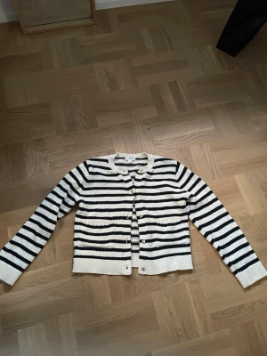 Randig kofta med guldknappar - Säljer en snygg kofta från Gina Tricot med svartvita horisontella ränder. Koftan har rund halsringning, långa ärmar och knäppning framtill med guldiga knappar. Perfekt för en stilren och klassisk look.