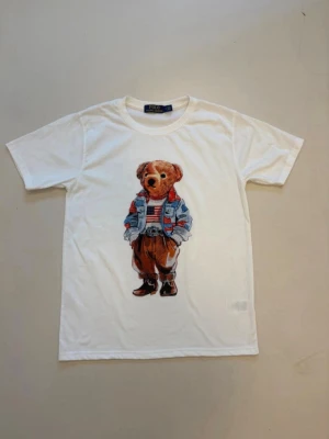 Vit t-shirt med Polo Bear från Polo Ralph Lauren - Vit t-shirt från Polo Ralph Lauren med ikoniskt Polo Bear-tryck framtill. Björnen har på sig jeansjacka, randig tröja och bruna byxor. Klassisk passform och rund halsringning. Perfekt för en avslappnad och cool stil.