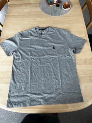 Grå t-shirt från Polo Ralph Lauren - Klassisk grå t-shirt från Polo Ralph Lauren med broderad svart logga på bröstet. T-shirten har rund halsringning och korta ärmar, perfekt för en clean och stilren look.