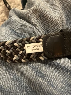 Flätat bälte från ONEWEAR Sweden 100cm - Säljer ett snyggt flätat bälte från ONEWEAR Sweden i mörka och ljusa toner. Bältet har detaljer i skinn och ett klassiskt spänne i silver. Perfekt för att ge din outfit en cool touch.