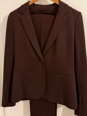 Brun kavaj och byxa kostym - Snygg brun kostym med klassisk kavaj och matchande byxa. Kavajen har smal passform, enkel knappstängning och två fickor framtill. Materialet känns mjukt och följsamt, perfekt för en stilren look.