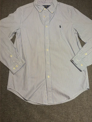 Randig skjorta från Ralph Lauren - Säljer en klassisk blåvit randig skjorta från Ralph Lauren med knappar framtill och långärmad design. Skjortan har en snygg krage och ikonisk logga broderad på bröstet. Perfekt för dig som gillar stilrena plagg med premiumkänsla.