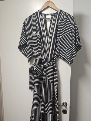 Svartvit jumpsuit med grafiskt mönster - Cool svartvit jumpsuit med geometriskt mönster och vida ben. Djup v-ringning framtill, korta ärmar och knytskärp i midjan. Materialet känns mjukt och flowy, perfekt för dig som gillar att sticka ut med stilrena detaljer.