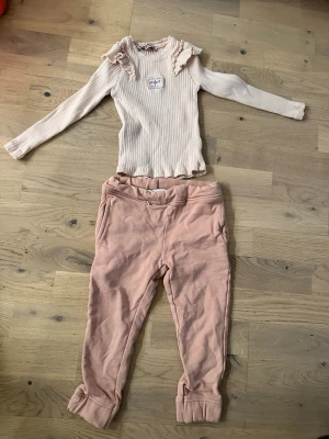 Ljusrosa loungewear-set från Newbie - Ljusrosa loungewear-set från Newbie Mysigt loungewear-set från Newbie i ljusrosa nyanser. Setet består av en ribbad långärmad topp med volangdetaljer vid axlarna och en liten etikett framtill, samt mjuka byxor med resår i midjan och uppvikta benslut. Perfekt för chill dagar hemma.