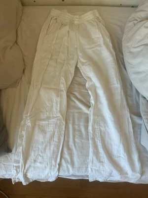 Vita linnebyxor med resårmidja - Säljer ett par vita linnebyxor med bred resår i midjan och raka ben. Byxorna har sidfickor och är tillverkade i ett lätt och luftigt linnematerial som är perfekt för varma dagar. De har en avslappnad passform och enkel design.
