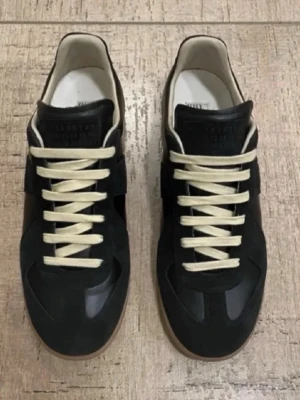 Svarta  Maison Margiela gats - Säljer ett par svarta sneakers från Maison Margiela med beigea snören och bruna gummisulor. Skorna har en klassisk låg profil och är tillverkade i en mix av läder och mocka. Perfekta för dig som vill ha en stilren och trendig look.