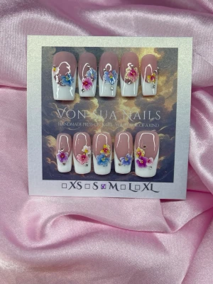 Flora (M);Medium Square - Handgjorda press-on naglar i storlek M: 16 mm, 12 mm, 12,5 mm, 11,5 mm, 10 mm. Naglarna har en nuderosa bas med vit handmålad fransk tipp och dekorerade med små kristaller, fina-blommor i blått, rosa, gult och lila samt pärlor. Formen är fyrkantig och medium långa.