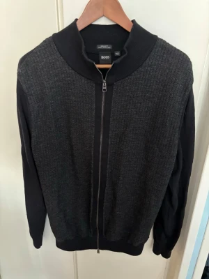 Tröja från Hugo BOSS - Mörkgrå ziptröja från Hugo BOSS i ull med ribbad krage och strukturstickat mönster. Tröjan har hel dragkedja framtill. 