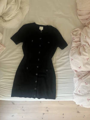Svart kort klänning med knappar - Svart kort klänning från H&M med rund halsringning och korta ärmar. Klänningen har dekorativa knappar längs hela framsidan och två fickor framtill. Materialet är mjukt och stretchigt, vilket ger en bekväm passform.