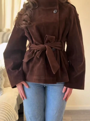 Mörkbrun trenchcoat med knytskärp Gina tricot slutsåld  - Snygg mörkbrun trenchcoat med hög krage, knappar framtill och ett knytskärp i midjan. Jackan har vida ärmar och axelklaffar. Jackan är oanvänd och prislappen är kvar. 