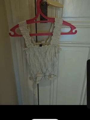 Beige rynkad linne med breda axelband - Säljer ett beige linne med volangprydda axelband och rynkad struktur. Linnet har en stretchig passform och är tillverkad i ett mjukt tyg, perfekt för att skapa en söt och trendig look. Inga synliga loggor eller märken.