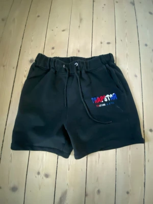 Svarta Trapstar shorts med snörning - Svarta shorts från Trapstar med broderad logga i blått, vitt och rött på sidan. De har elastisk midja med snörning och är tillverkade i mjukt material, perfekt för en avslappnad look.