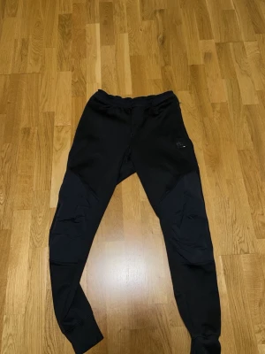 Svarta mjukisbyxor från Nike - Svarta mjukisbyxor från Nike med slim fit och ribbade muddar vid benslut. Byxorna har diskret Nike-logga på framsidan och dragkedja på bakfickan. Materialet är mjukt och stretchigt, perfekt för chill eller träning.
