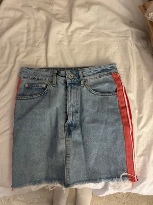 Jeanskjol med röda ränder - Säljer en ljusblå jeanskjol med rå kant och röda sidoremsor med vita ränder. Kjolen har klassisk femficksdesign och dragkedja framtill. Perfekt för dig som gillar sportig och avslappnad stil. 