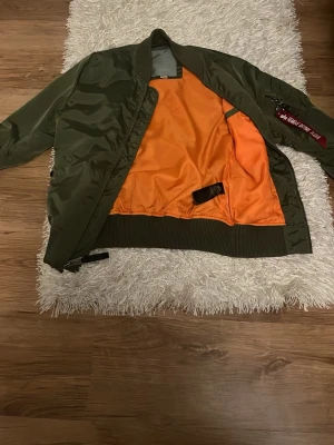 Grön bomberjacka från Alpha Industries - Snygg grön bomberjacka från Alpha Industries med klassisk orange insida och ribbade muddar. Jackan har en ficka med dragkedja och röd tag på ärmen samt två sidofickor. Tillverkad i glansigt nylonmaterial för en cool look.