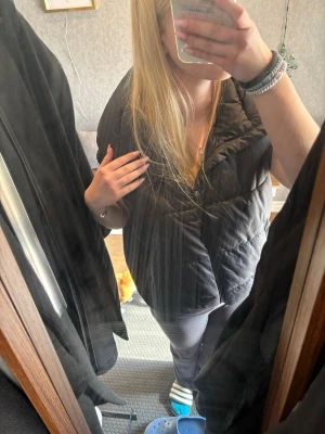  Svart dunväst från 157 - Säljer en svart dunväst från 157 med dragkedja framtill. Västen har en puffig look och är tillverkad i ett glansigt material som ger en modern vibe. Perfekt att slänga över en hoodie eller tröja för extra värme.  