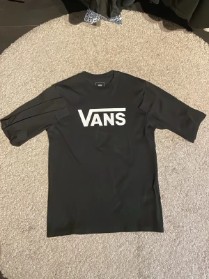 Svart Vans t-shirt med logga - Svart t-shirt från Vans med klassisk vit logga på bröstet. T-shirten har rund hals och långa ärmar, perfekt för en avslappnad streetstyle-look. Materialet är mjukt och känns skönt mot huden. 14+ år stl XL