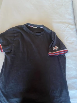 Svart Moncler t-shirt med logga - Säljer en svart t-shirt från Moncler med korta ärmar och snygga röd, vit och blå detaljer på ärmslut och halsringning. Moncler-loggan sitter på ena ärmen och materialet är mjuk bomull. Perfekt för dig som gillar stilrena plagg med exklusiv känsla.