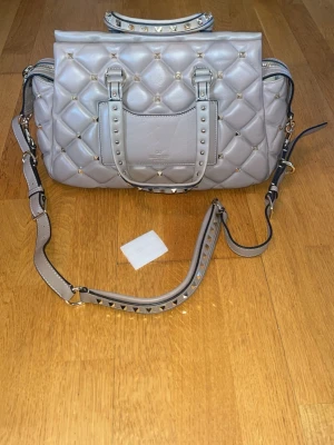 Valentino väska beige (ny skick) - Säljer en beige quiltad handväska från Valentino med guldfärgade nitar och detaljer. Väskan har både handtag och justerbar axelrem, samt en ficka framtill. Tillverkad i läder och har en cool, edgy look med sina pyramidformade nitar. 100% äkta, ny skick.