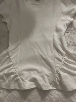 Vit topp med rynkade detaljer - Säljer en vit topp med korta ärmar och snygga rynkade detaljer på sidorna. T-shirten har en figurnära passform och är gjord i mjukt bomullsmaterial. Perfekt för dig som gillar en enkel men trendig look.