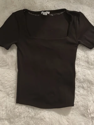 Mörkbrun topp med fyrkantig hals - Säljer en mörkbrun topp från Newyorker med fyrkantig halsringning och korta ärmar. Toppens passform är slim och den är gjord i mjuk bomull med lite stretch. Perfekt att styla med jeans eller kjol för en clean look.