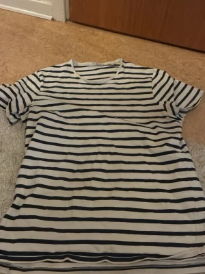 Randig t-shirt från Bläck - Säljer en vit t-shirt från Bläck med svarta horisontella ränder. Klassisk rund halsringning och korta ärmar. Perfekt för dig som gillar en enkel och stilren look. Materialet är mjukt och känns skönt mot huden.