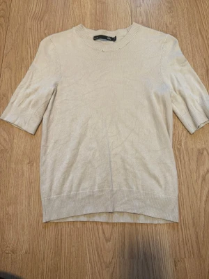 Beige stickad topp från Lager 157 - Säljer beige stickad kortärmad topp från lager 157 med rund hals och korta ärmar. Enkel och stilren designe. Materialet är mjukt och skönt, och toppen har ribbade detaljer vid hals, ärmslut och nederkant.