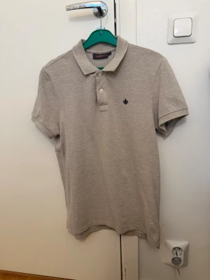 Beige pikétröja med knappar - Klassisk beige pikétröja från Morris Stockholm med korta ärmar, krage och två knappar framtill. Tröjan har en diskret broderad detalj på bröstet och är gjord i mjuk bomull för en clean och enkel stil. Pris kan diskuteras