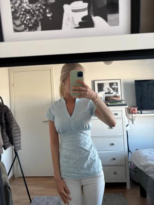 Ljusblå blus med v-ringning - Säljer en ljusblå blus med v-ringning och korta ärmar. Blusen har en figurnära passform och knappar framtill. Materialet ser ut att vara lätt och bekvämt, perfekt för en avslappnad stil. Diskret randigt mönster ger en snygg touch.