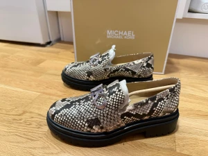 Ormmönstrade loafers Michael Kors - Snygga loafers från Michael Kors med ormmönstrad ovandel i beige, svart och grå toner. Skorna har chunky svart sula och en dekorativ metallogo på ovansidan. Tillverkade i konstläder med trendig design som sticker ut.