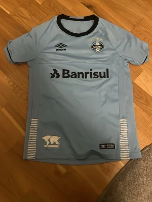 Ljusblå Grêmio matchtröja från Umbro - Snygg ljusblå matchtröja från Grêmio med svarta detaljer och klubbmärke på bröstet. Tröjan har tryckt sponsorlogga 'Banrisul' och vita ränder på sidorna. Tillverkad i lätt och ventilerande material, perfekt för fotboll eller träning.